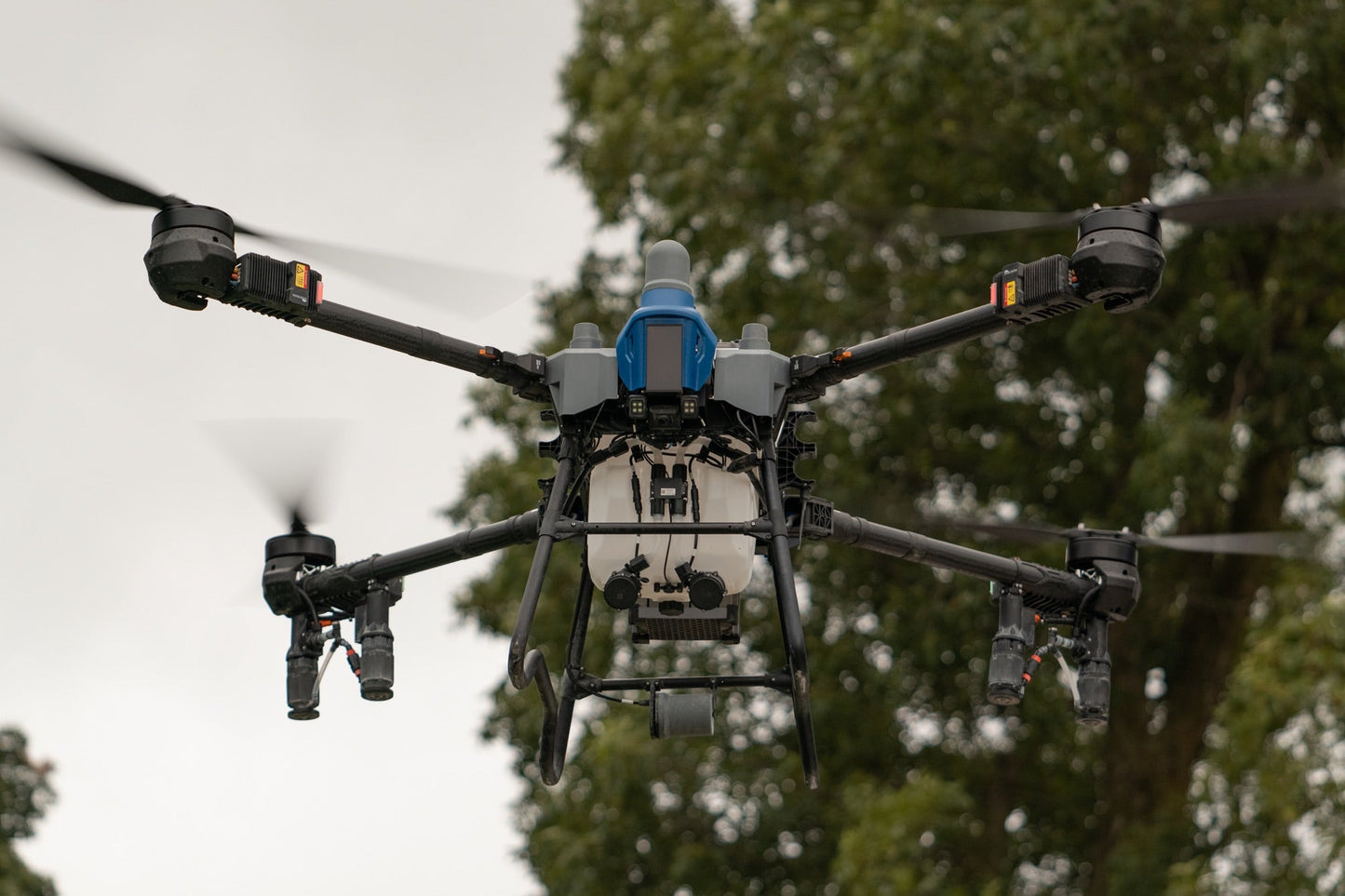 EAVision J150 spray drone hovering with rotors turning, spray nozzles and booms visible beneath the airframe and the liquid tank in the centre