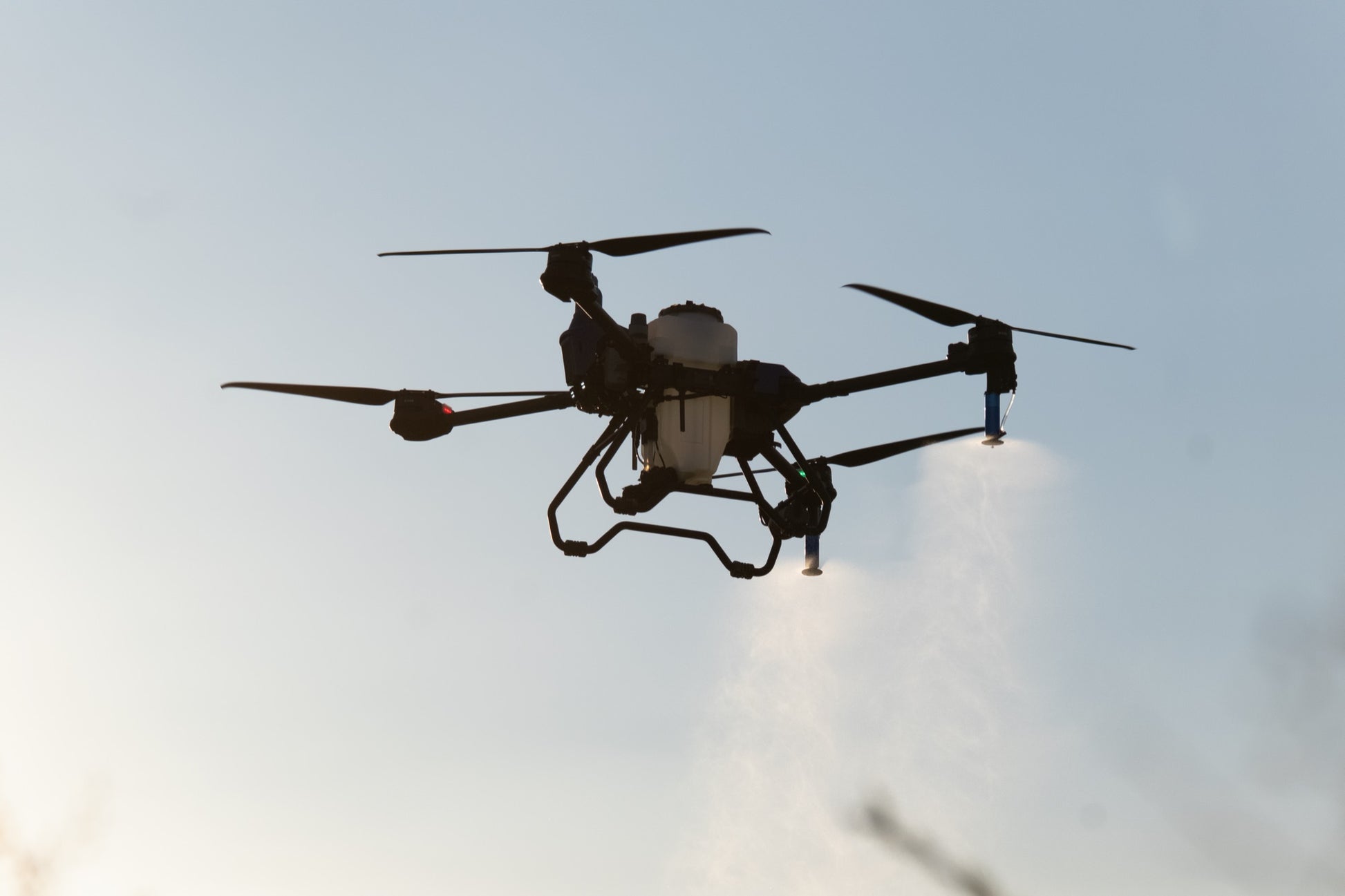 EAVision J70 spray drone in flight against a bright sky, four rotors turning and spray nozzles running
