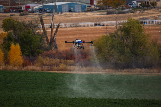 EAVision J70 spray drone applying product low over a green field, spray mist visible beneath the booms with farm buildings behind
