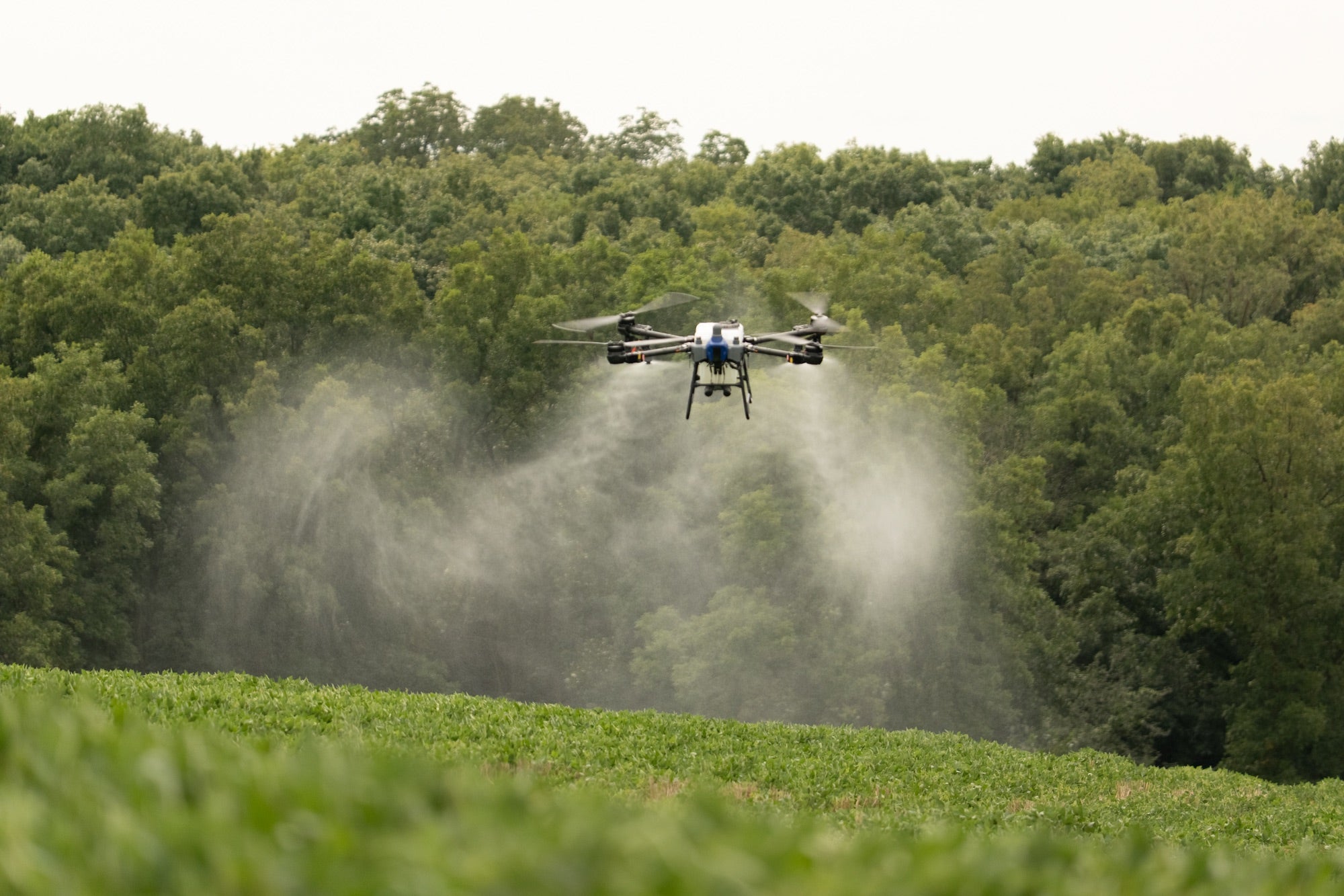 EAVision J150 spray drone applying fungicide over an Illinois soybean field, spray plume fanning out beneath the booms against a treeline